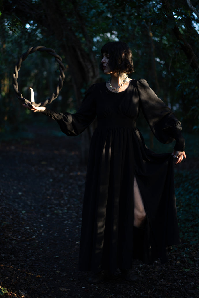 A witch in the woods at sunset with a long black dress and a clay portal candle holder with a lit candle