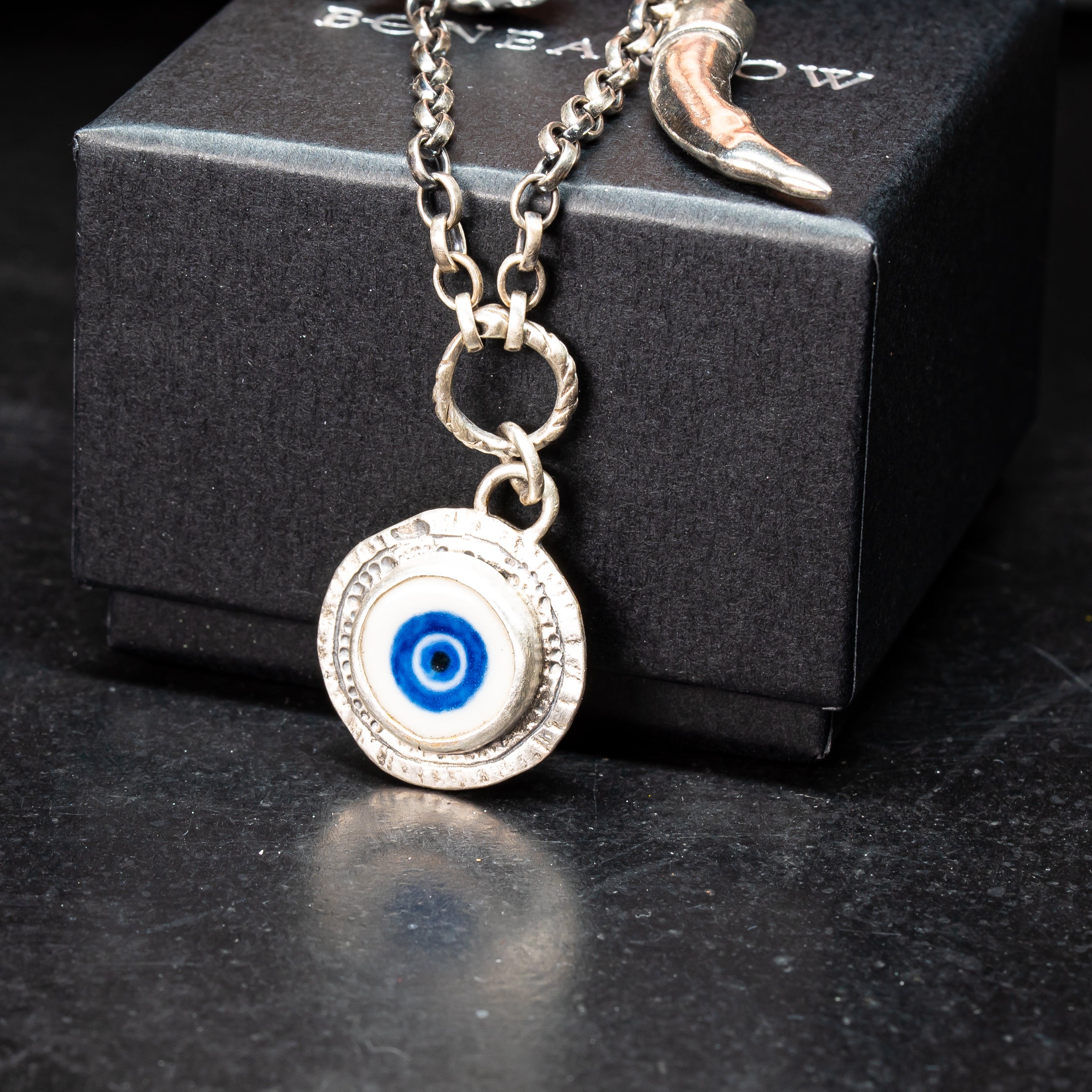 porcelain handprinted evil eye motif set in a sterling silver handmade coin pendant on handmade charm enclave draped over a jewellery box on a black background
