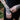 A close-up of a person's tattooed hands and wrist adjusting a silver chain bracelet on their left wrist, wearing a black lace dress outdoors with blurred foliage in the background.
